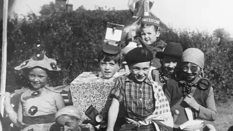 Great Dunmow Carnival A black and white photo of several children wearing fun costumes, including one as a pirate. There is a hedgerow behind them.