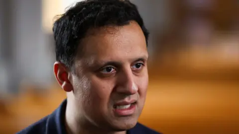 Anas Sarwar has dark hair and is wearing a dark t-shirt