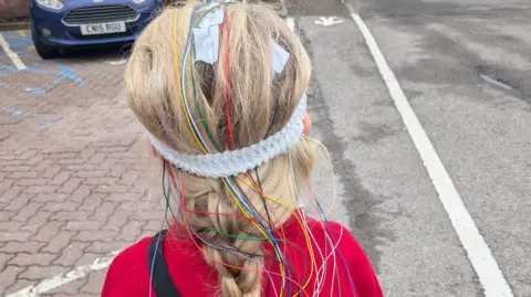 Emma Alderman The behind of Noa-Rose's head which has many multi-coloured wires coming out of. She is stood in a car park. 