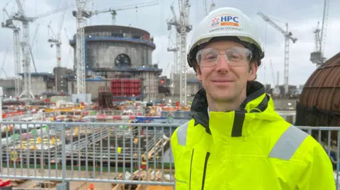 Andrew Cockroft from Hinkley Point C stands in a hi-vis yellow jacket and hard hat in front of the huge building site at Hinkley Point. Numerous cranes and the concrete reactor building are behind him