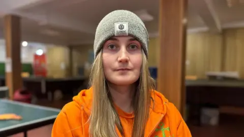 Teaghan Daly wears a neon orange hoodie and grey hat. She has long blonde hair and its in an empty room with columns and a table tennis table behind her