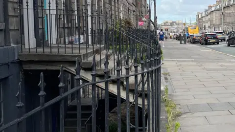 Railings marking the area of a basement. There is a pavement with steps rising up to a green doorway above. there are cars on the street.