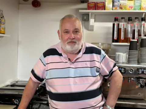 Mark Breen with a grey beard, wearing a pink striped polo shirt standing in his cafe kitchen with mugs and disposable cups seen behind him