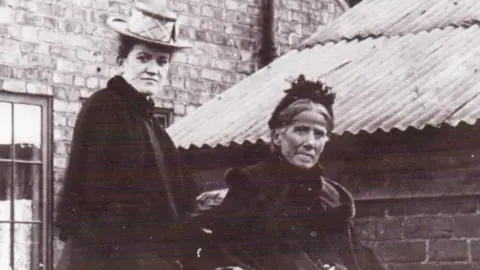 Heanor & District Local History Society A black and white archive image of two women dressed in clothing of the late 1800's. The younger woman stands to the left with short dark hair, a rimmed hat and a black cloak. The older woman is sat to the right, with fairer hair tied back, a decorative dark hat and thick dark coat. She is seated in a wheelchair. Behind the pair are brick buildings with a tin roof.
