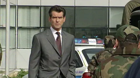 Pierce Brosnan stands outside an office building. He is wearing a grey suit and has thick, dark brown hair. He is frowning slightly and has a hand in his pockets. There are people in camoflauge facing away from the camera and an emergency car which says 'Polis' on it.