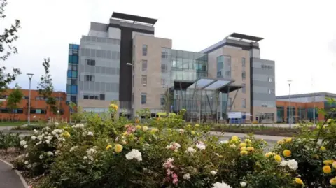 NHS Front and side aspect of Peterborough City Hospital. The reception area is in view. In the foreground is a rose bush. The building is brown coloured with extensive glass frontage.