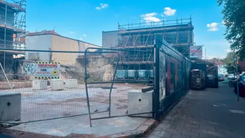 An empty plot of land on the corner of a residential and high street with harris fencing and billboards