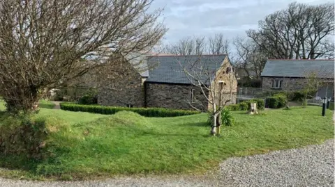 An existing guest room at the Plume of Feathers, the building is made of brown brick and has grey tiles on the roof. Around the building is grass and small trees. 