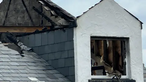 A close-up image of the second floor and roof space of Sixty Six. The roof has been burnt away, with exposed concrete brick and a steel beam remaining in place along with grey slate on the rood. There is also a burnt out window, with damage to the upstairs of the building.