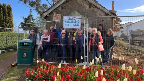 Protesters at the Furzeham toilet block in Brixham. They are stood behind a temporary metal fence. They are holding up signs which say 'Save Brixhams Heritage' and 'Don't rock the block'. There are tulips in front of the fence. Behind the protesters is a derelict building.