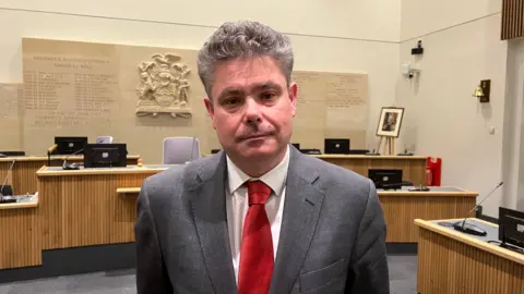 Simon Dedman/BBC John Kent is wearing a white shirt, red tie and gray suit jacket. He has brown hair. He is standing in the council chamber.