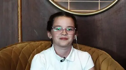 A young girl wearing glasses and a white polo shirt sitting in a mustard coloured chair. She is looking into the camera.