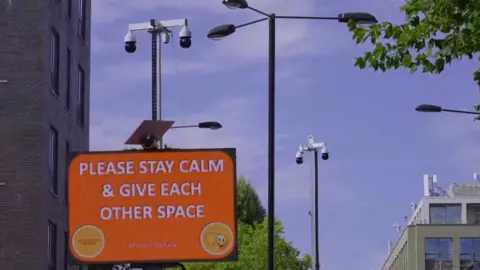 An orange sign with the words please stay calm and give each other space attached to a lamp post with CCTV cameras on top of it against a blue sky