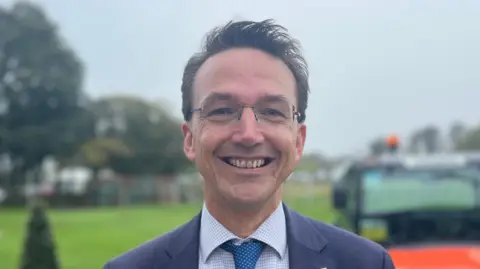 Deputy Kirsten Morel in a blue and white suite wearing glasses. He is smilling and short hair. THe background of the image is blurred.