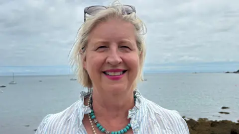 Karen Shenton-Stone is standing outdoors on a rocky shoreline overlooking St Catherine's Bay. She's wearing a light-blue and white striped shirt, layered necklaces including turquoise beads, and sunglasses resting on top of her head. In the background, several small boats are anchored on the water, with rocky coastline visible along the edge of the bay. The sky is overcast with thick grey clouds.