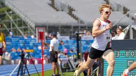 Onyeka Okoh A young man wearing a sports jersey and black shorts. He had the number 89 on his top and is wearing sunglasses. Behind him is a stadium and he is running on a blue running track. 