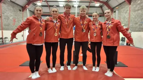 Helen Duggan Six gymnasts stand on a red mat, they wear red jackets, black leggings and white socks. They have their arms around each other and all wear medals on colourful ribbons. 