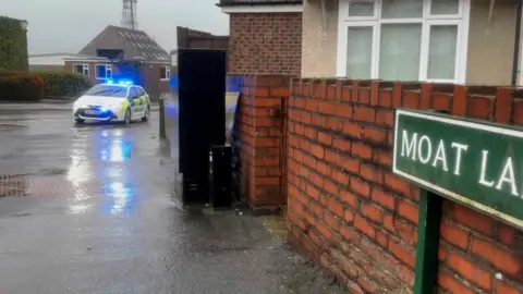 A rainy picture with puddles on. The road sign "Moat Lane" is partly visible and a police car with a blue light can be seen. 