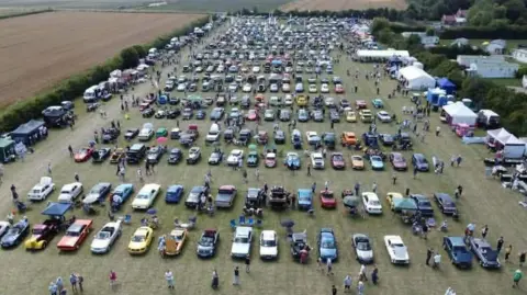 Aerial view of a previous car show at the site. There are hundreds of vehicles parked in rows on a field, with stands around the perimeter. There are static caravans in a nearby field.