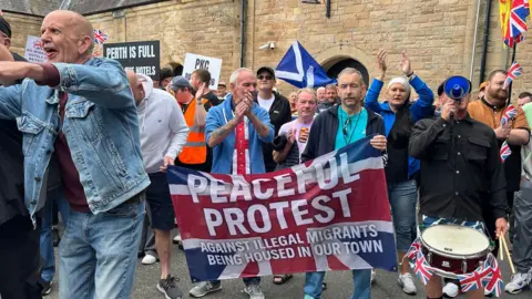 A group of people clapping and holding signs which say things like "perth is full" and "peaceful protest against illegal migrants being housed in our town"