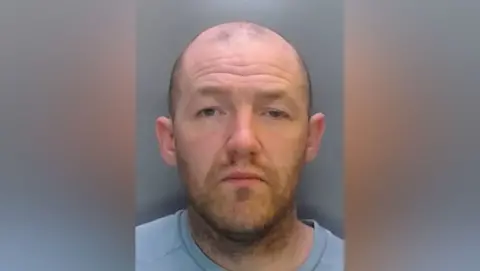 Mugshot of Cliff. He is balding with very short dark hair and a stubbly ginger beard