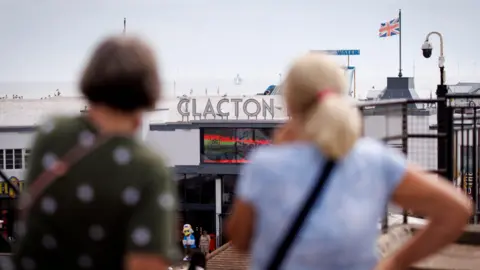 EPA/Shutterstock Two women looking towards Clacton Pier on a grey day. The pier has "Clacton" in large letters and a Union Jack flag above its entrance.