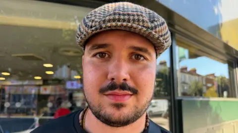 Richard Knights/BBC Zak Apsey is wearing a checkered cap and a dark T-shirt in Clacton.