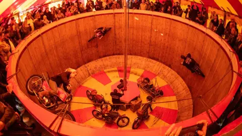 Ken Fox This image shows a live stunt show where multiple motorcyclists ride around the vertical wooden walls of a circular arena, often called a “wall of death,” while a crowd watches from above. The riders move horizontally along the steep sides, using speed and balance to stay up, while several parked bikes sit at the bottom near a central pole.
