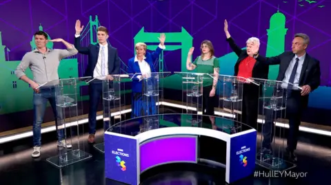 Luke Campbell and the five defeated mayoral candidates taking part in a BBC debate in a TV studio. The candidates stand in a semi-circle. All have their hands raised, except Campbell whose hand is half-raised.
