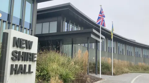 Brian Farmer/BBC A modern glass-fronted building with the words 'New Shire Hall' on a stone to the right. There are two flag poles, one flying the Union flag and the other the flag of Ukraine.