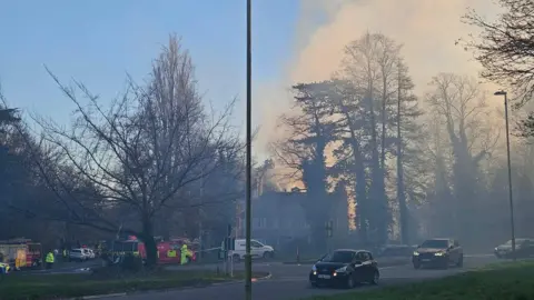 A hazy and smokey scene with cars in the foreground of a road where there are emergency services in the distance.