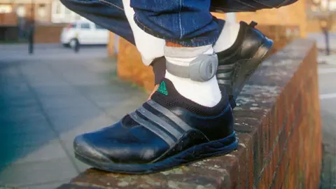 A man is wearing an ankle monitoring tag. We can only see his black trainers, white socks and bottom of his blue jeans. He is perched on top of a waist-high wall.