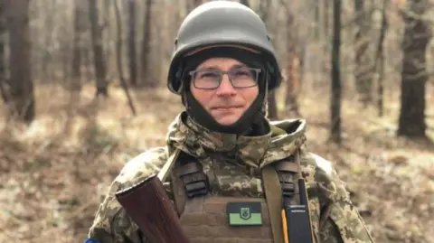 Dmytro A man dressed in combat gear stands in a wooded area. He looks directly at the camera. He is wearing an army hard hat and glasses. You can see the top of a gun he is holding.