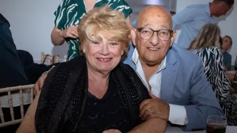 Elaine has short blonde hair and wears a navy blue dress with a scarf around her shoulders. Philip has his arm around her and is wearing a light grey blazer and white shirt