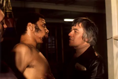 A scene from Play For Today : Gangsters from 1975 showing bare-chested actor Paul Barber on the left with an unidentified male actor wearing a beige polo necked jumper and leather jacket 

