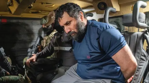Drug trafficker Adolfo Macias Villamar, aka 'Fito', is seen ub grey trousers and a blue T-shirt, as hehe is moved by armed soldiers in Guayaquil, Ecuador, on 25 June 2025.
His hands are handcuffed behind his back. He has a black beard and black hair. The soldiers are armed and are wearing balaclavas to conceal their identities. 