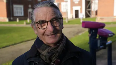 John Fairhall/BBC Stuart Grimwade, standing outside, wearing a black coat and scarf, he has on glasses and short grey hair. He has a building behind him and binoculars on two stands with purple tops. 