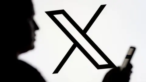 Man looks at X app on a smartphone with the platform's logo displayed in the background.