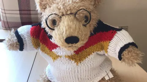 Stuart Race A teddy bear wearing a knitted white shirt with black, red and gold patterns around the chest and shoulders to match Germany's football kit. The teddy bear is also wearing glasses.