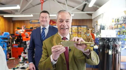 PA Media Reform UK leader Nigel Farage holds a key for '10 Downing Street', as he and Reform UK mayoral candidate Ryan Coogan, standing behind him, visit a key cutting shop