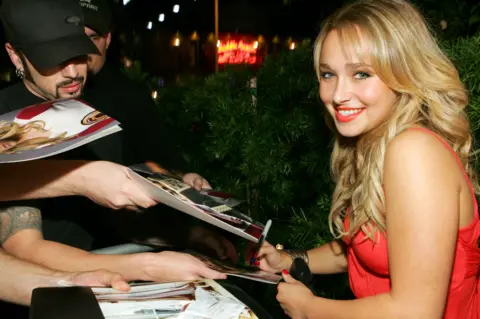 Getty Images Hayden Panettiere during Teen Vogue Young Hollywood Party on the red carpet signing autographs. She is wearing a red dress and smiling at the camera 
