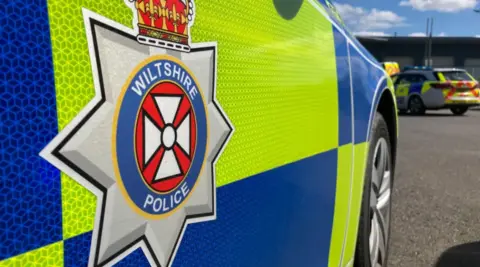 Close up shot of a police car which says 'Wiltshire Police' on the side. There are other police vehicles in the background.