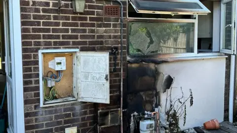 A burnt section of a wall beneath a window. There is a gas meter nearby and another meter on the ground beneath the burnt area