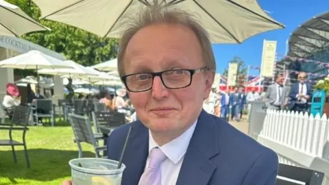 A man with short blonde/grey hair sits in a pub garden with tables, chairs and garden umbrellas behind him. There is a small white fence behind him and other people walking along a path. He is wearing black-rimmed glasses, a blue suit, white shirt and lilac tie. He is holding a clear plastic glass with a drink, slice of lemon and black straw in. 