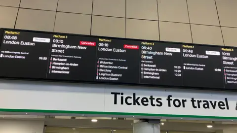 A train station with an information board with train times on it, showing cancellations and information. The screen is black and white. 
