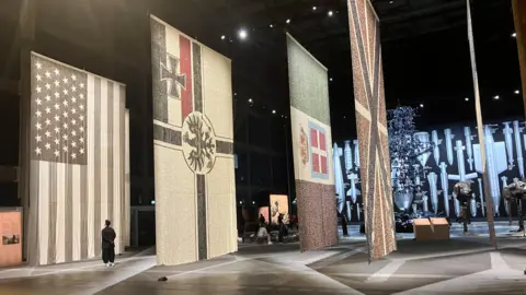 Photograph of a series of flags at Manchester's Aviva Studios. The flags were made with hundreds of thousands of buttons.
