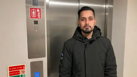 BBC A man with dark hair and a beard wearing a black coat, standing in front of a set of lift doors and looking straight at the camera. On the wall next to him is a little screen with a picture of a spanner on it. 