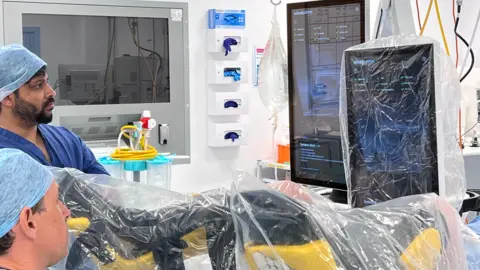 A surgeon and a colleague watch large medical monitors beside equipment covered with sterile plastic sheeting during a procedure in an operating theatre.