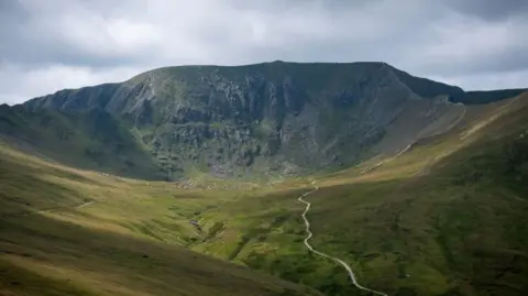 A look towards Helvellyn mountain in The Lake District. A narrow path leads towards the summit, which is surrounded by a large green field.