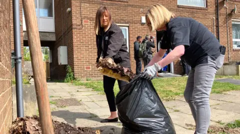 Nicola Rees/BBC Two women shovelling rubbish into a black bin bag on a housing estate